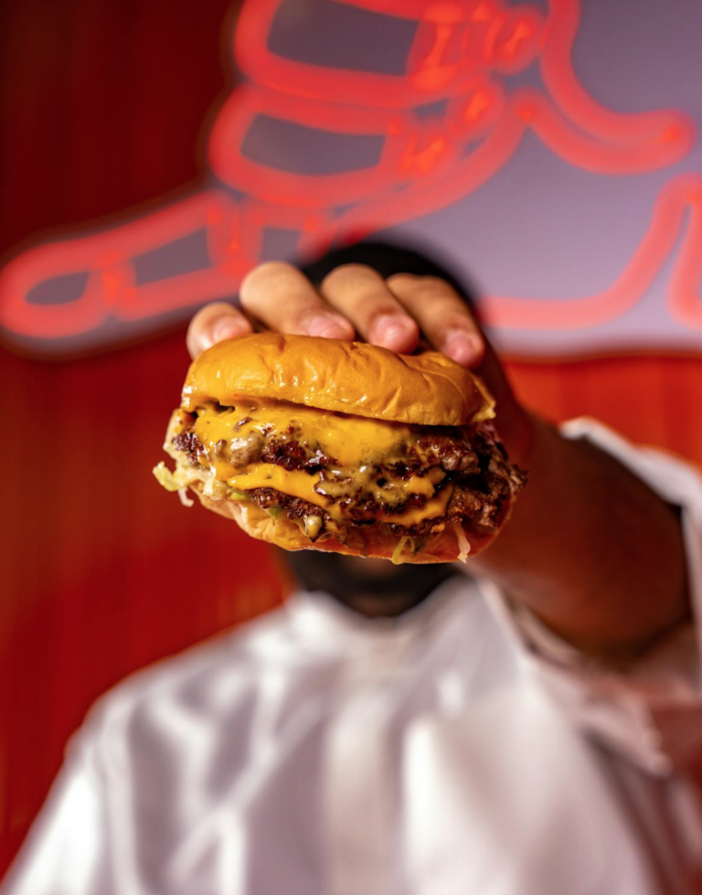Kok holder en dobbelt cheeseburger frem foran rødt neonlys